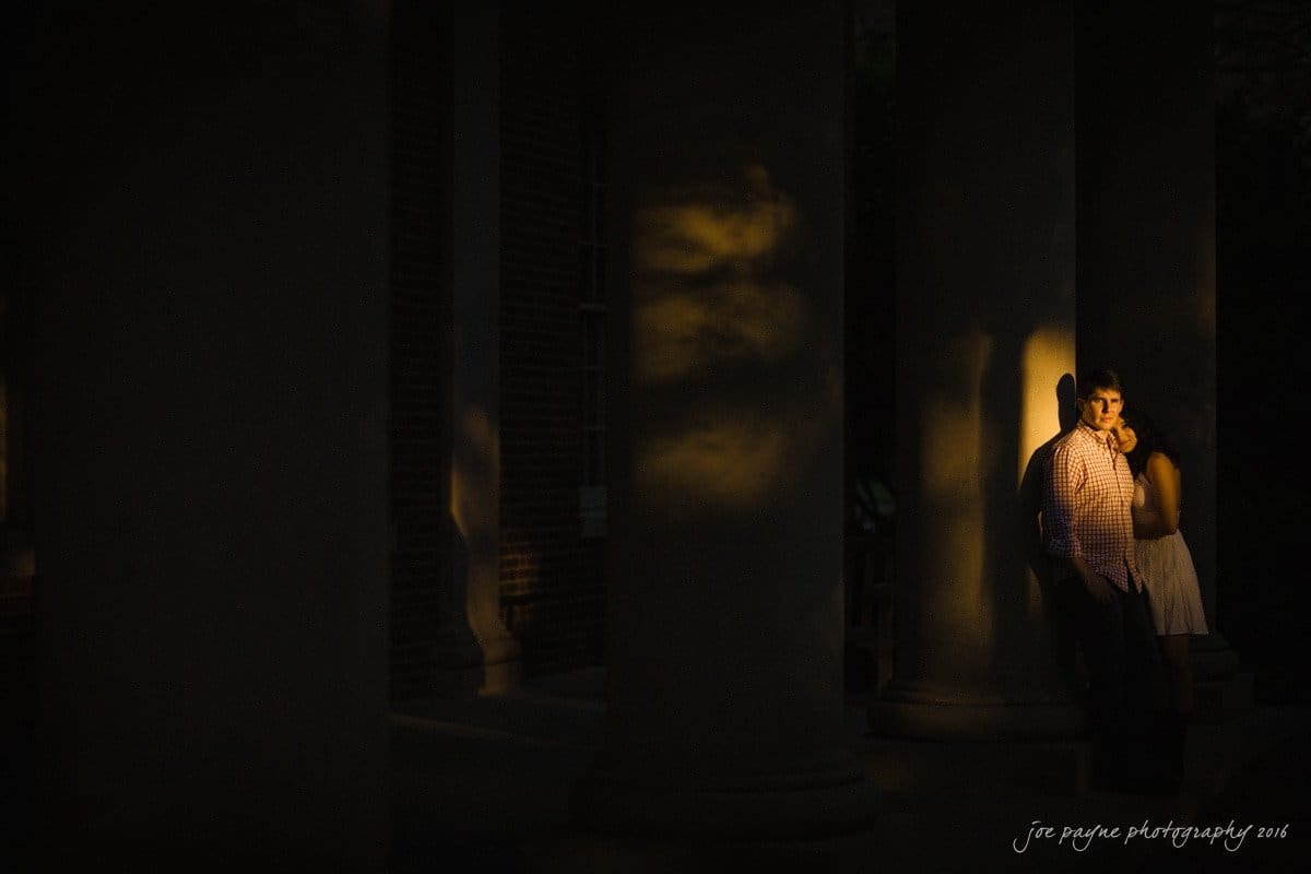 A couple stands in dim lighting between large columns. The man leans against a column, wearing a plaid shirt, while the woman stands beside him in a light dress. Soft light casts shadows around them, creating a serene, intimate atmosphere.