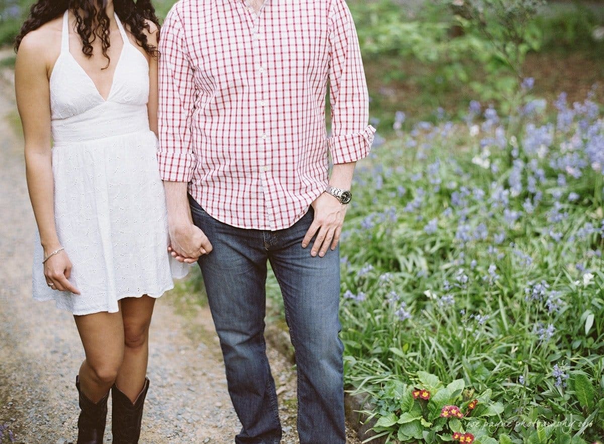 A couple holding hands on a path lined with wildflowers. The woman is wearing a white dress and black boots, while the man is in a checkered shirt and jeans.