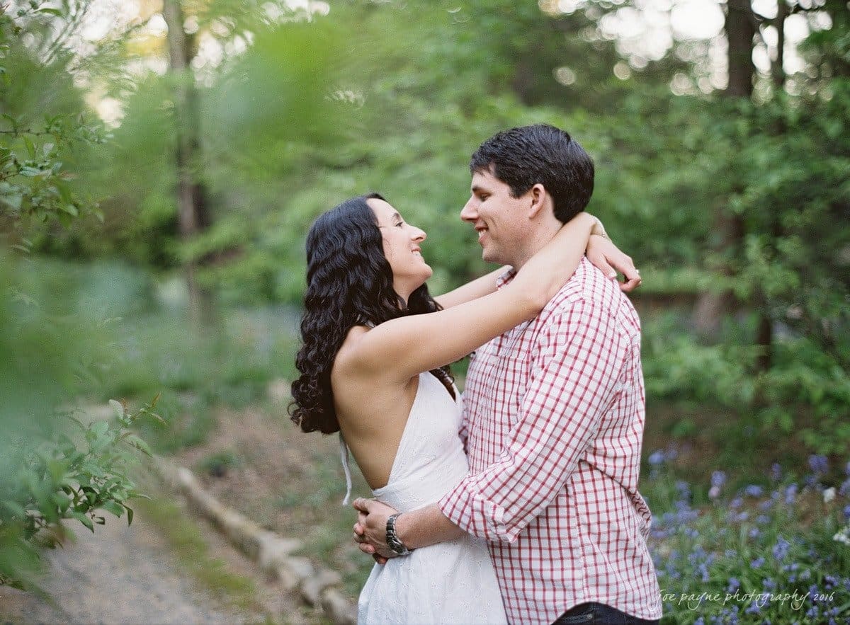 A couple embraces and smiles at each other in a lush garden setting. The woman is wearing a white dress, and the man is wearing a red checkered shirt. Greenery surrounds them, creating a serene and romantic atmosphere.