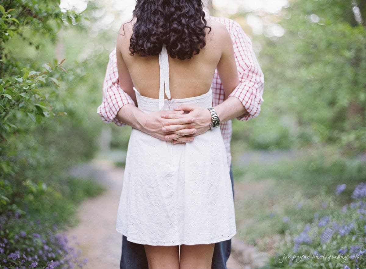 A couple embracing outdoors on a path surrounded by greenery and purple flowers. The woman, wearing a white dress, faces away from the camera while the man in a plaid shirt wraps his arms around her waist.