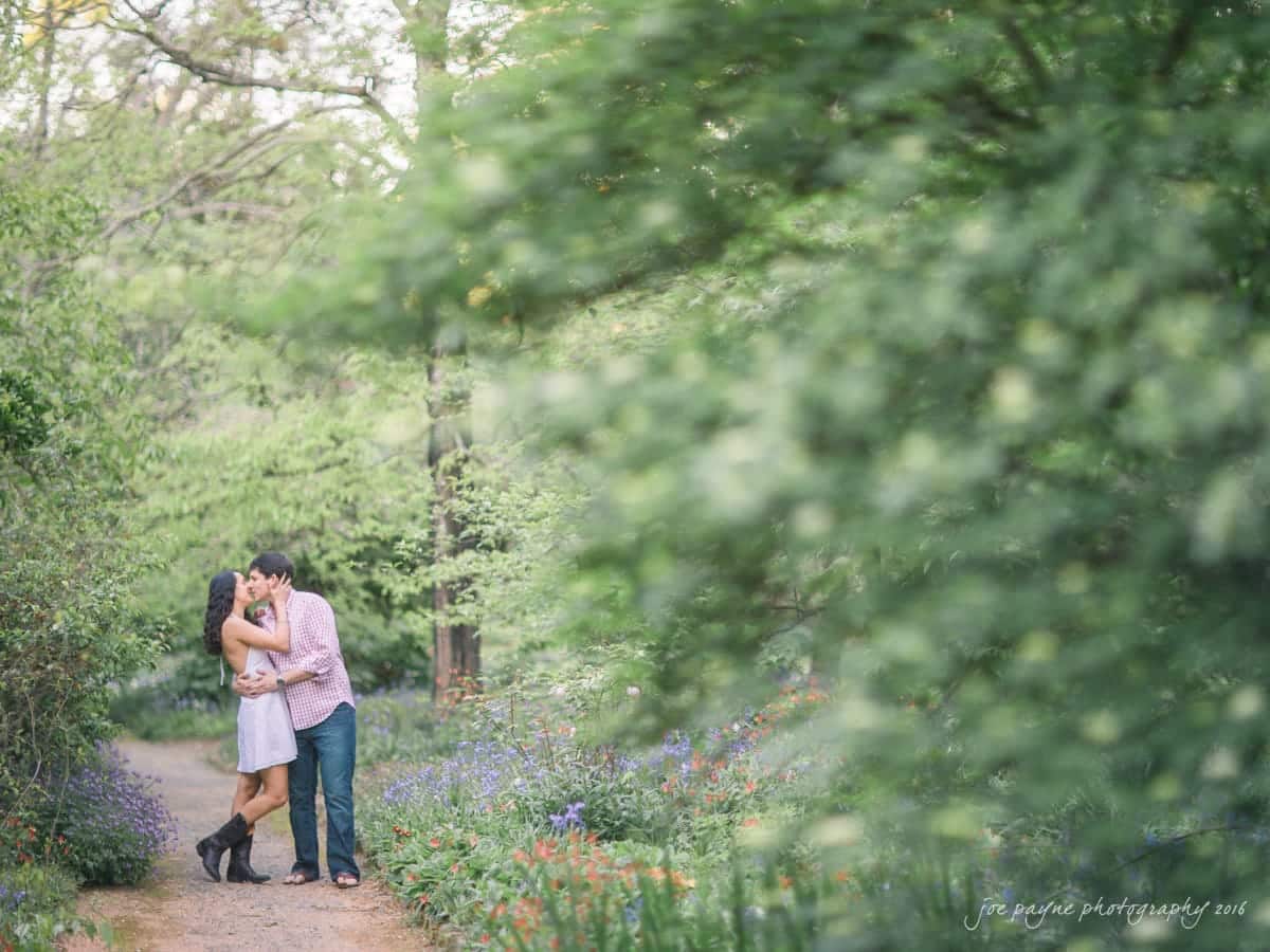 A couple embraces on a narrow path surrounded by lush greenery and colorful wildflowers. The scene is serene and romantic, with soft, natural lighting filtering through the trees.