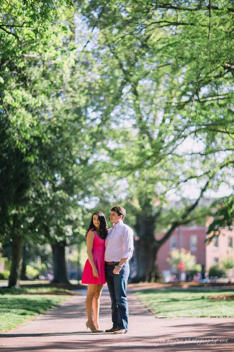 A couple stands on a sunlit path in a park, surrounded by lush green trees. The woman wears a pink dress, and the man is in a white shirt and jeans. They look off into the distance on a bright, clear day.