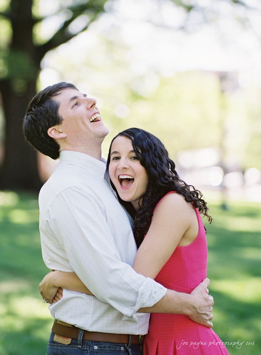 A couple is embracing and laughing joyfully in a sunlit park. The man is wearing a white shirt, and the woman is in a pink dress. The background is blurred, showing green grass and trees.