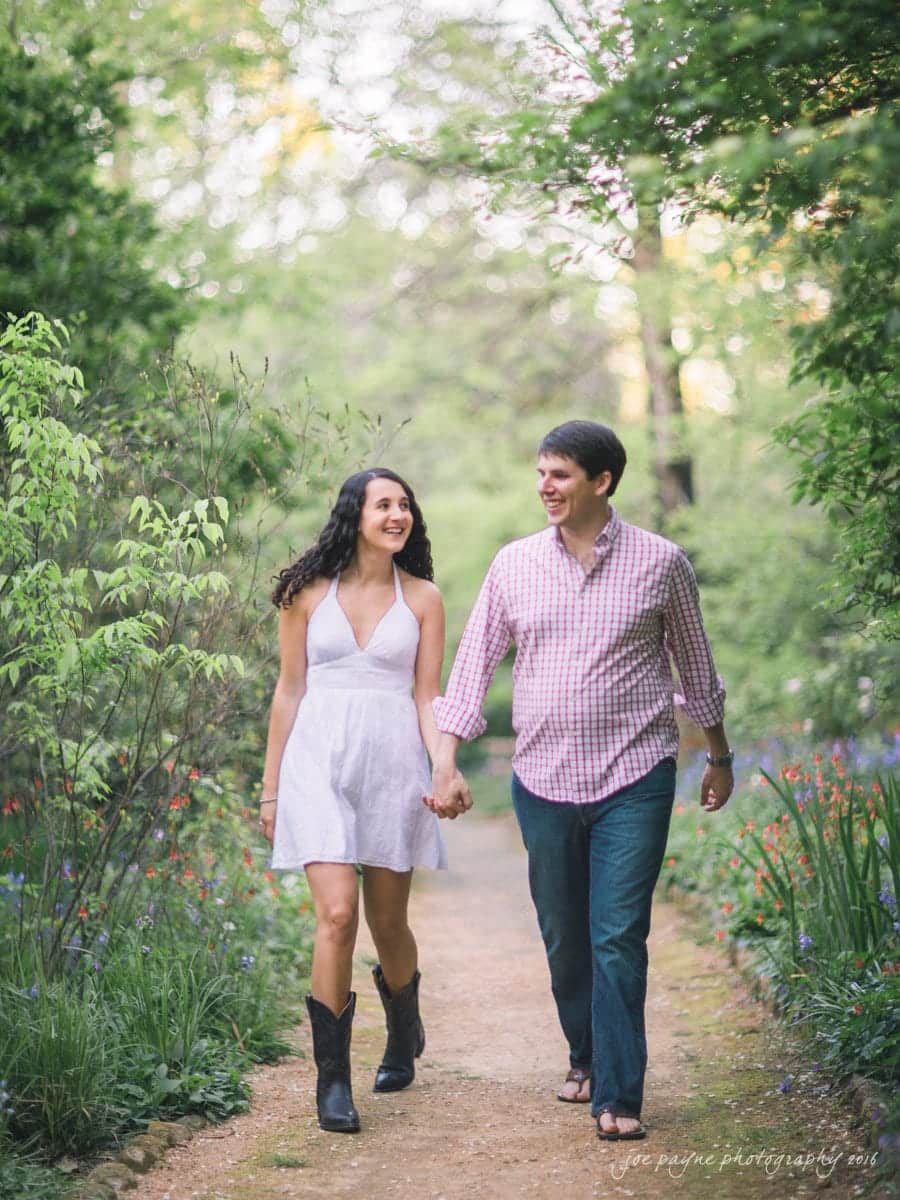 A couple walks hand in hand down a garden path. The woman is wearing a white dress with black boots, and the man is in a pink checked shirt and jeans. They are surrounded by lush greenery and flowers, smiling at each other.