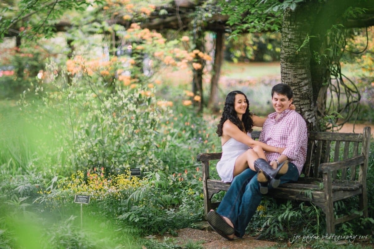A couple sits on a wooden bench in a lush garden, surrounded by vibrant flowers and greenery. The woman is wearing a white dress, and the man is in a checkered shirt and jeans. They appear relaxed and happy, enjoying each others company.