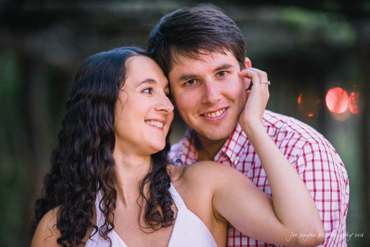A smiling couple poses closely outdoors. The woman, with long curly hair, touches the mans face. The man, wearing a checkered shirt, looks at the camera while the woman gazes at him fondly. They are surrounded by a blurred natural background.