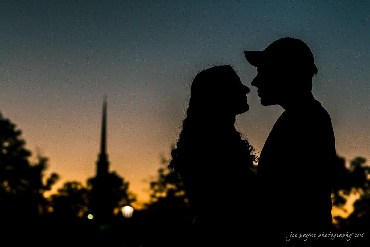 Silhouetted couple facing each other in front of a sunset, with tree outlines and a church steeple in the background. The sky transitions from orange to blue.