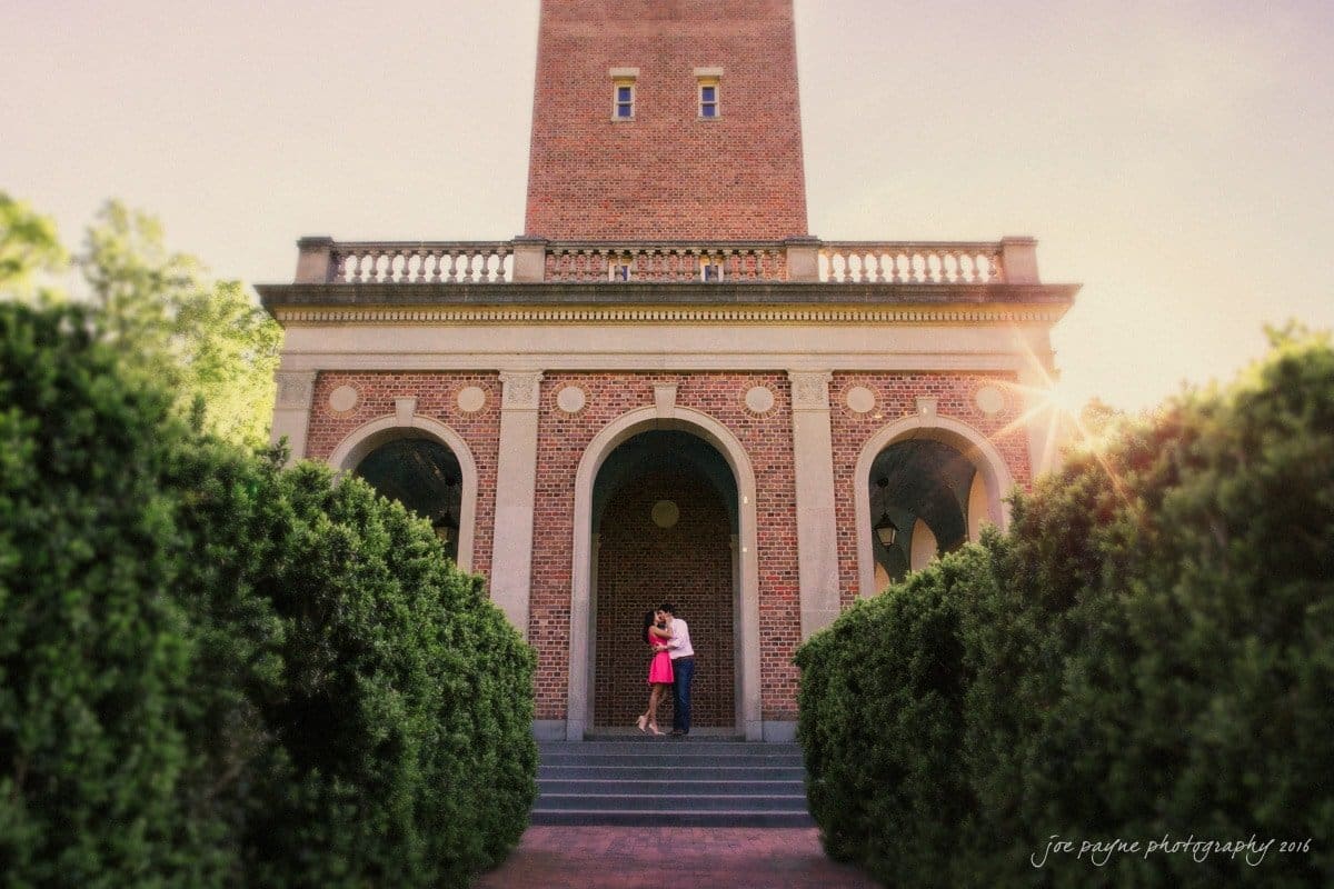 A couple embraces in front of a historic brick building with arched doorways. Tall hedges frame the walkway leading to them. The sun peeks through, creating a warm glow across the scene.