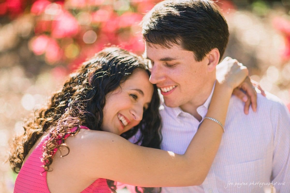 A couple joyfully embraces, with the woman smiling and leaning her head on the mans shoulder. The background features vibrant pink flowers in soft focus, creating a warm and intimate atmosphere.