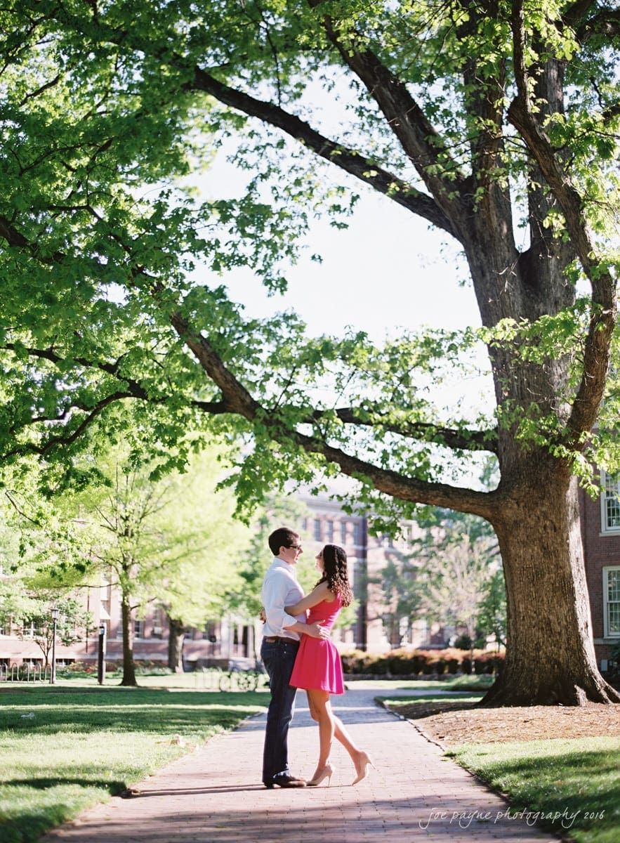 A couple embraces on a sunlit pathway under a large tree with lush green leaves. The woman is wearing a pink dress, and the man is in a white shirt and jeans. A brick building is visible in the background.