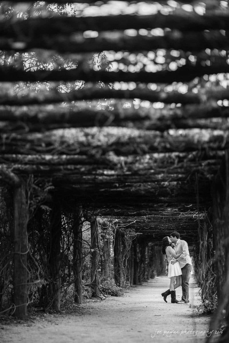 A black and white photo of a couple embracing under a wooden pergola covered with vines. The pathway is surrounded by nature, creating a romantic atmosphere. The couple stands in the center, holding hands and smiling at each other.