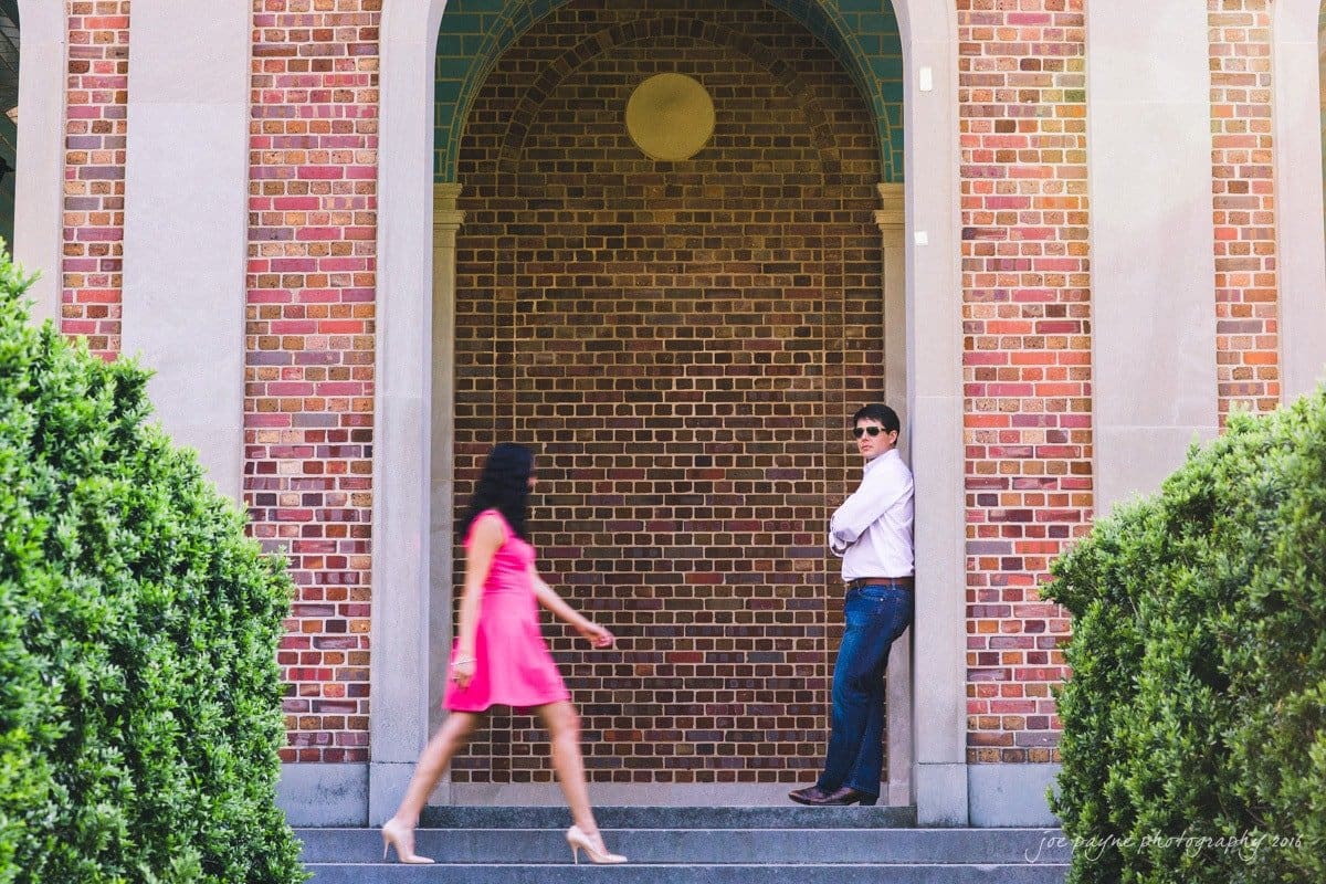 A woman in a bright pink dress walks past a man in a white shirt and jeans, who is leaning against a brick archway. Green shrubs frame the scene, adding a natural touch to the architectural setting.