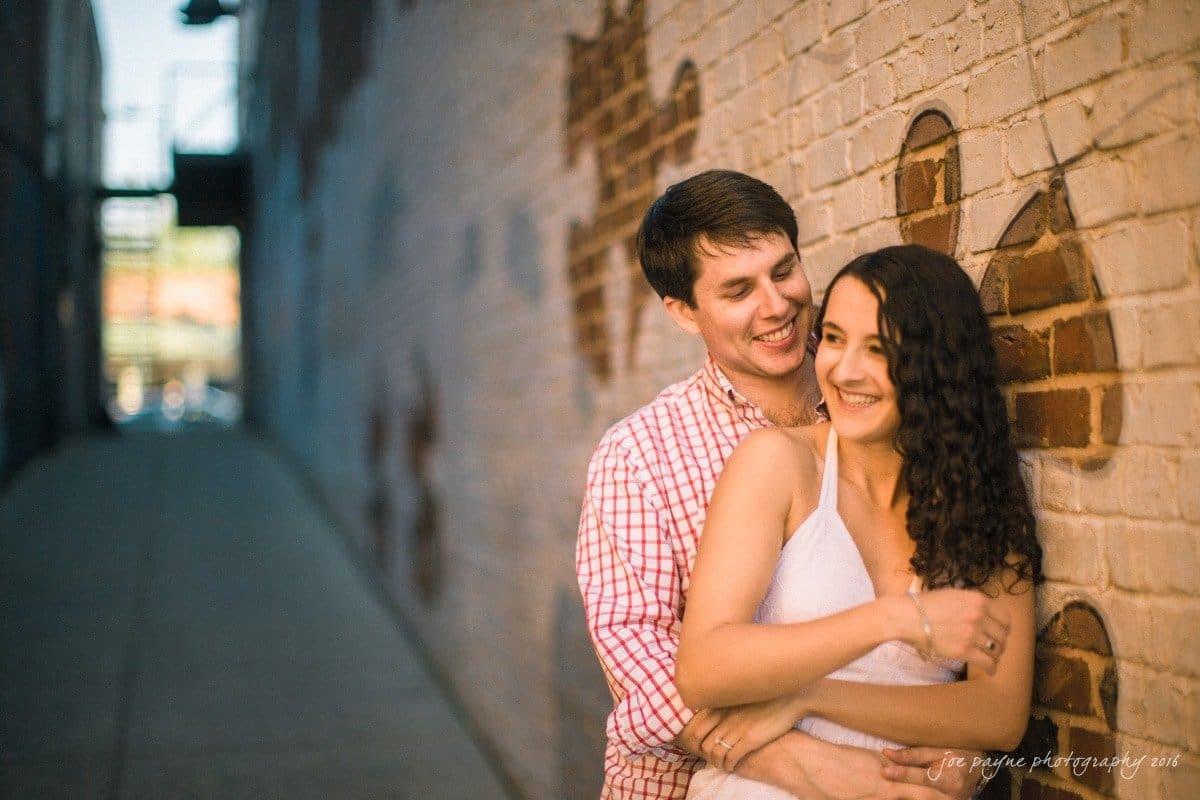 A couple embraces and laughs in an alley with a brick wall featuring heart-shaped designs. The man wears a red checkered shirt, and the woman is in a white dress. The alley is softly lit with a blurred background.