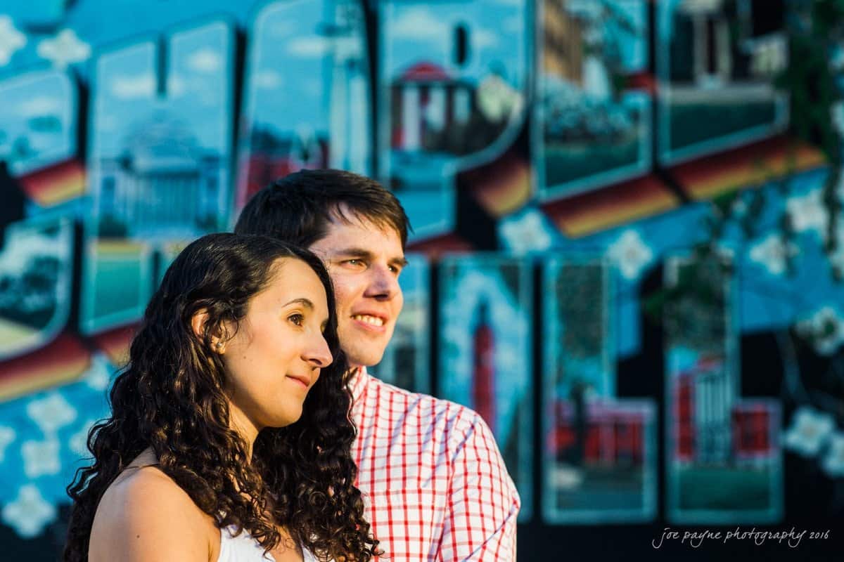 A couple stands side by side, gazing into the distance. The woman has long curly hair and wears a white top, while the man wears a red checkered shirt. In the background, there is a colorful mural with various images.