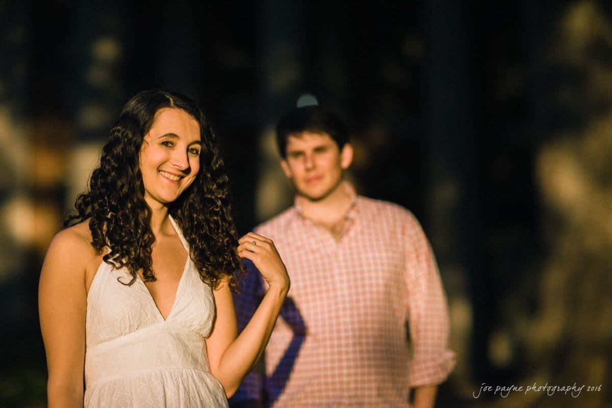A woman with curly hair in a white dress stands smiling in the foreground, while a man in a checkered shirt stands in the background, slightly out of focus, in a sunlit outdoor setting.