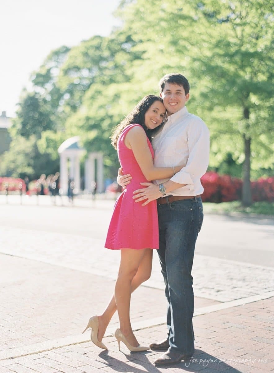 A couple stands happily together outdoors. The woman in a pink dress and heels leans against the man in a light button-up shirt and jeans. Sunlight filters through the trees in the background, creating a bright and joyful atmosphere.