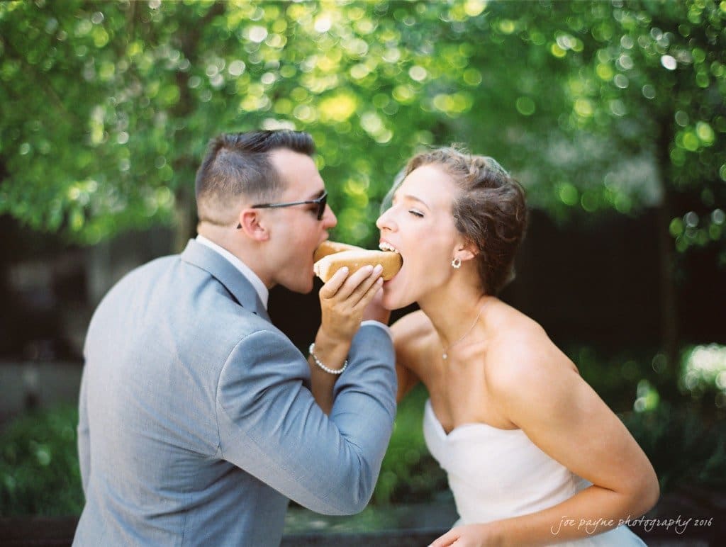 A couple in formal attire shares a bite from a large, shared sandwich outdoors. The man is wearing sunglasses and a suit, while the woman is in a white dress. Lush green trees create a soft, natural background.