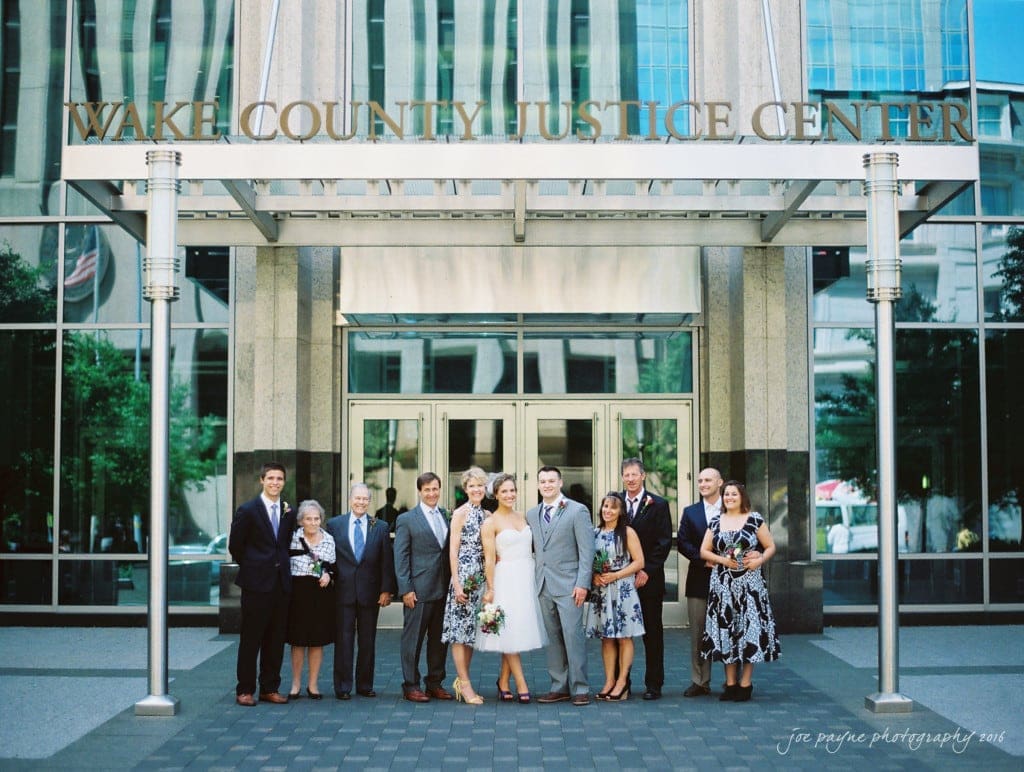 A group of people dressed in formal attire stand in front of the Wake County Justice Center. The building features a modern glass facade and signage above. The group appears to be posing for a photo on a sunny day.