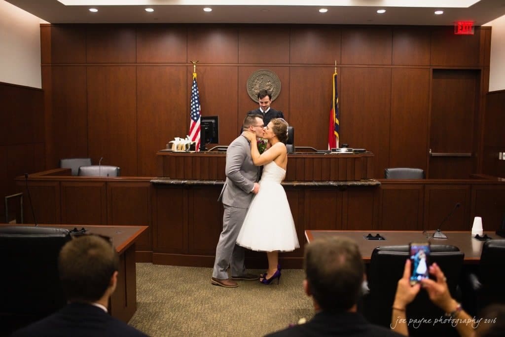 A couple in wedding attire shares a kiss in a courtroom setting. The officiant stands behind them, and there are flags and a seal on the wood-paneled wall. Onlookers in the foreground capture the moment on their phones.