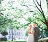 A bride and groom stand together in a sunlit park surrounded by lush green trees. The bride is in a white gown holding a bouquet, and the groom in a gray suit smiles beside her. A historic building is visible in the background.