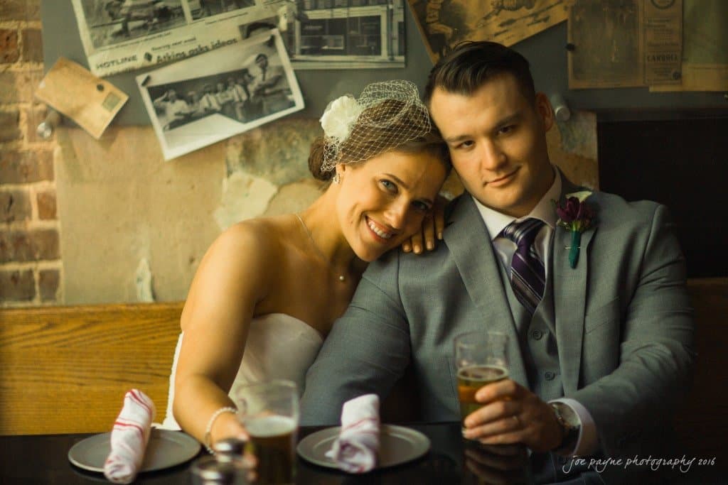 A couple in wedding attire sits together at a rustic restaurant table, each holding a beer. The woman, wearing a white dress and veil, rests her head on the mans shoulder. The man is in a gray suit with a purple tie. Decor includes vintage photos.