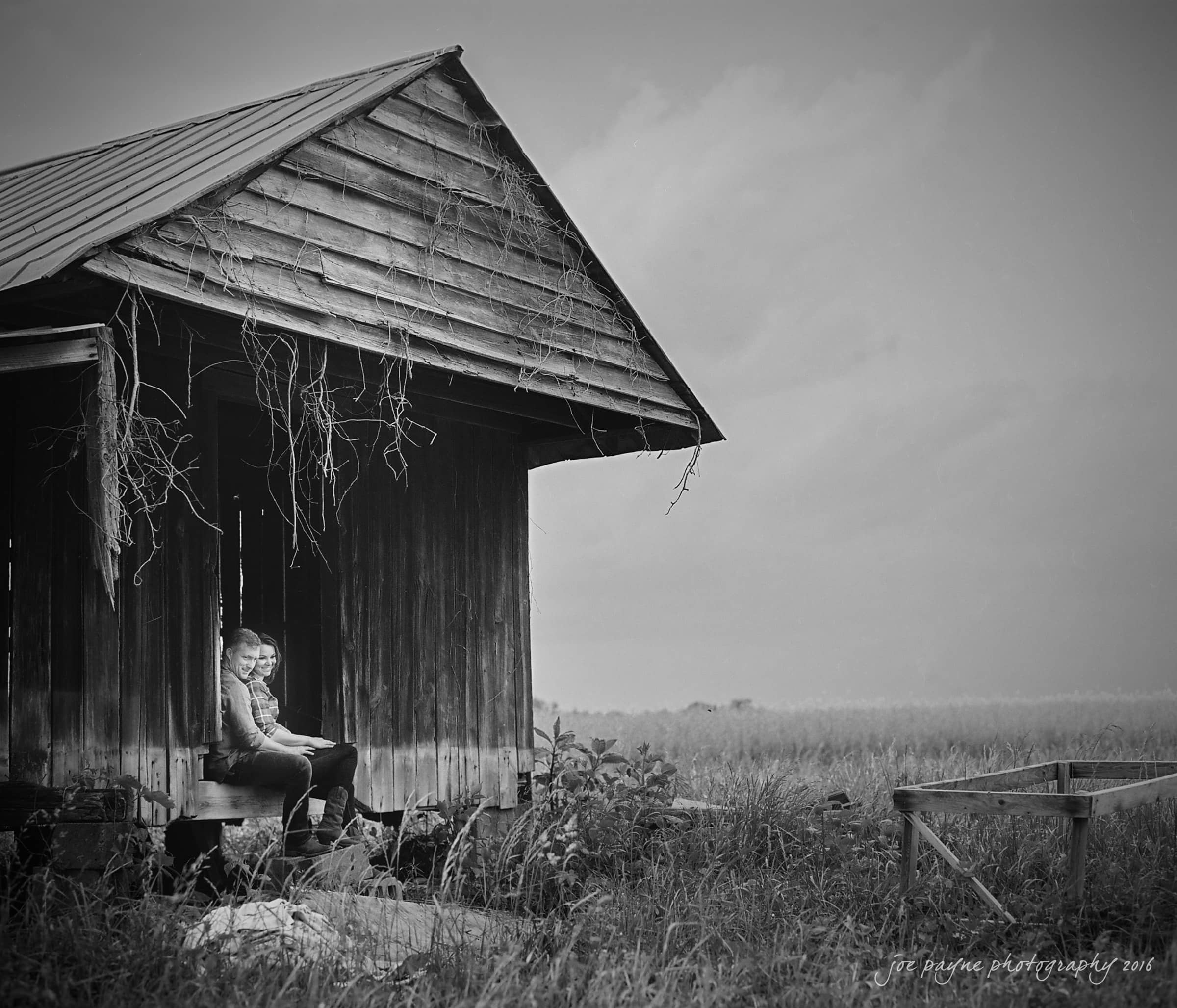 Black and white image of a person sitting on the steps of an old wooden shack with vines hanging from the roof. The shack is in a field under a cloudy sky, with an empty chair nearby.