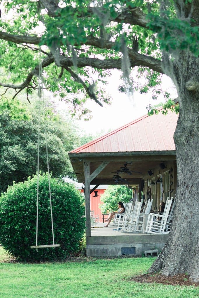 new bern wedding photographer - jessica & brinson A rustic porch with white rocking chairs sits under a red roof, surrounded by lush greenery. A tree with a hanging swing is in the foreground, casting shade on the grassy lawn. Moss drapes gently from the tree branches.