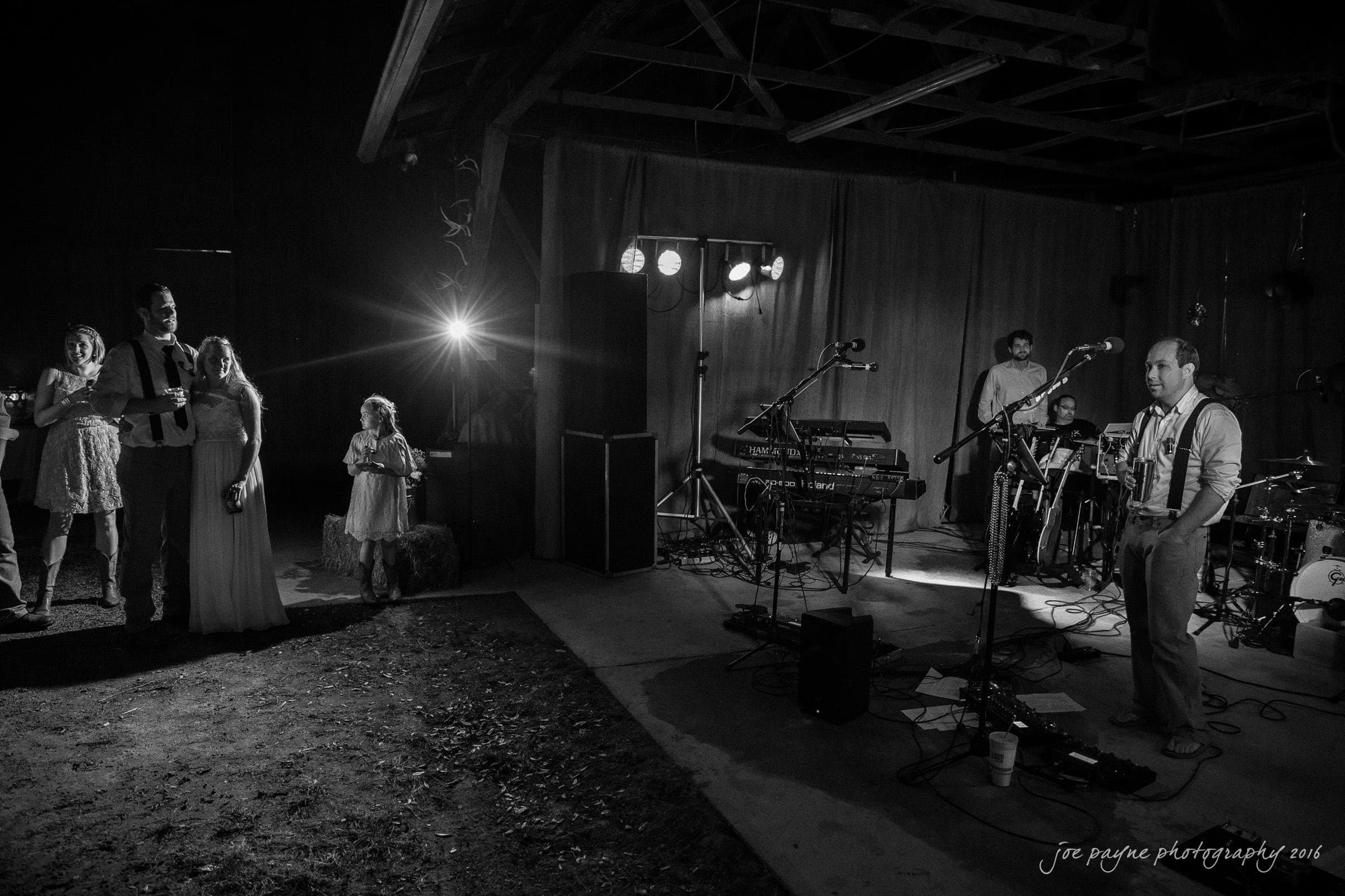 new bern wedding photographer - jessica & brinson A black and white photo of a band performing on a stage under a wooden canopy, with musical instruments and equipment. Several people, including a child and a couple, stand near the stage, illuminated by stage lights.