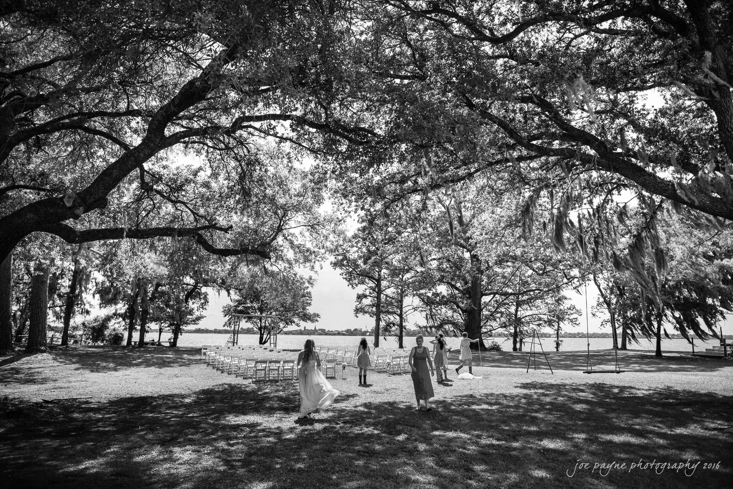 new bern wedding photographer - jessica & brinson A black and white photo of a serene outdoor wedding setting beneath large trees. Rows of chairs are arranged on the grass, overlooking a body of water. Children play nearby, and sunlight filters through the leaves.