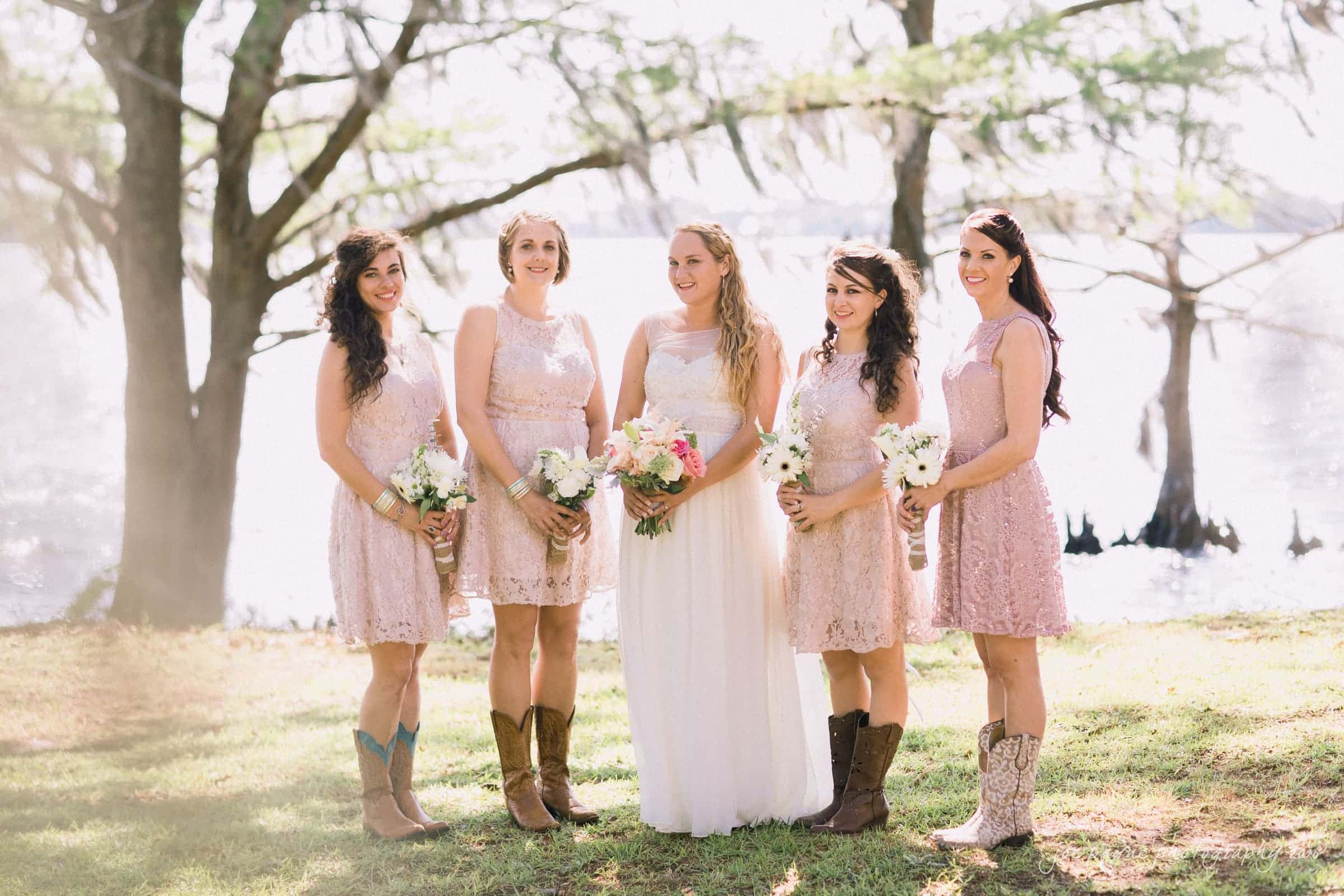 new bern wedding photographer - jessica & brinson A bride in a white dress stands with four bridesmaids in pale pink dresses, each holding bouquets. They are outdoors near a lake with trees in the background. Some of the bridesmaids are wearing cowboy boots.