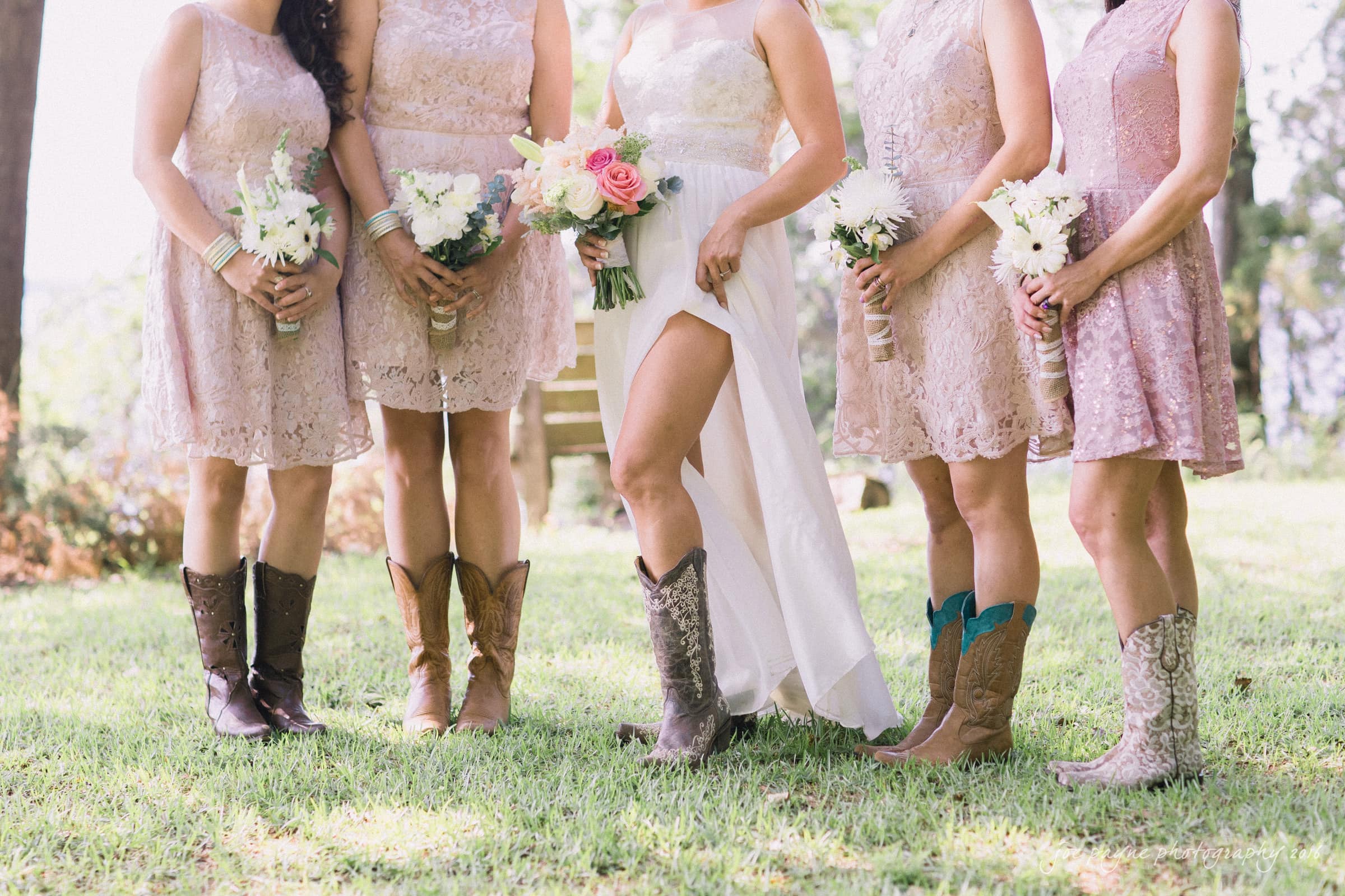 new bern wedding photographer - jessica & brinson A bride and four bridesmaids stand on grass wearing lace dresses and cowboy boots. Each woman holds a bouquet; the brides has pink and peach roses, while the bridesmaids have white flowers. They stand outdoors, likely at a wedding.