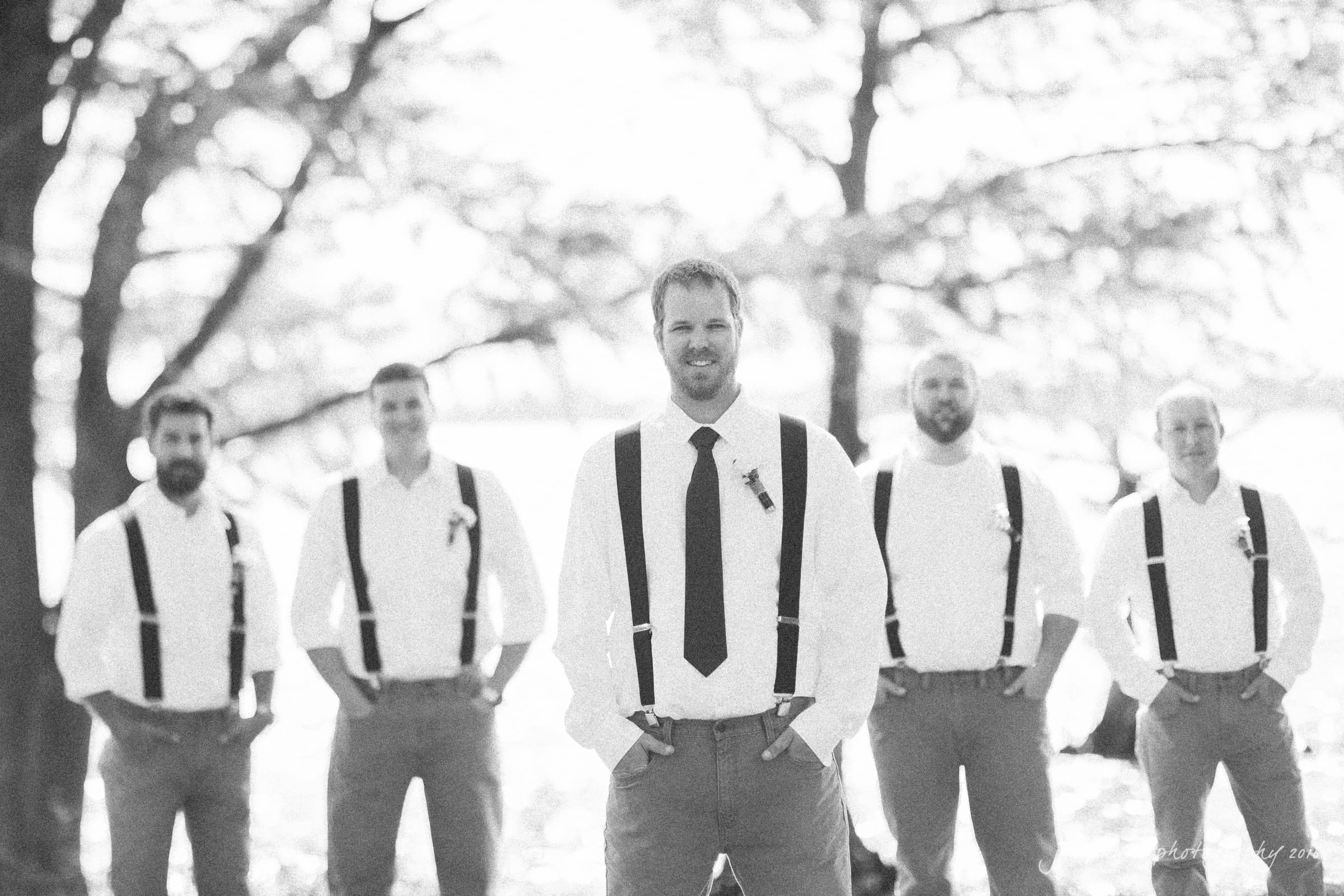 new bern wedding photographer - jessica & brinson A black and white photo of a group of five men wearing suspenders and white shirts, standing outdoors with trees and a body of water in the background. The man in the center has a tie, and they all appear to be posing for a formal occasion.