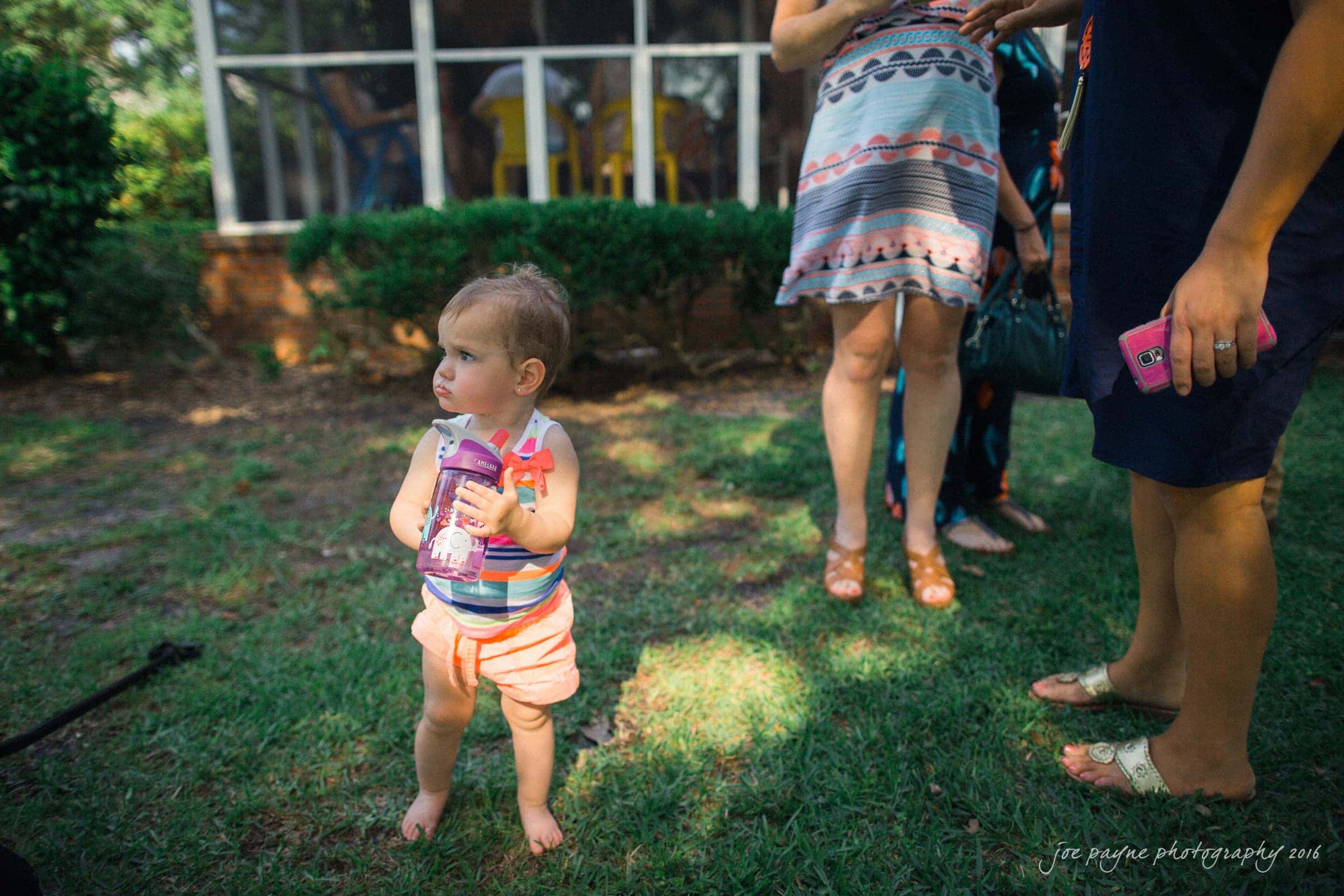 new bern wedding photographer - jessica & brinson A toddler in a striped outfit stands on grass, holding a purple sippy cup. Nearby adults, partially visible, wear casual summer clothing. A glass-enclosed area is in the background, along with some shrubs. Sunlight casts shadows on the scene.