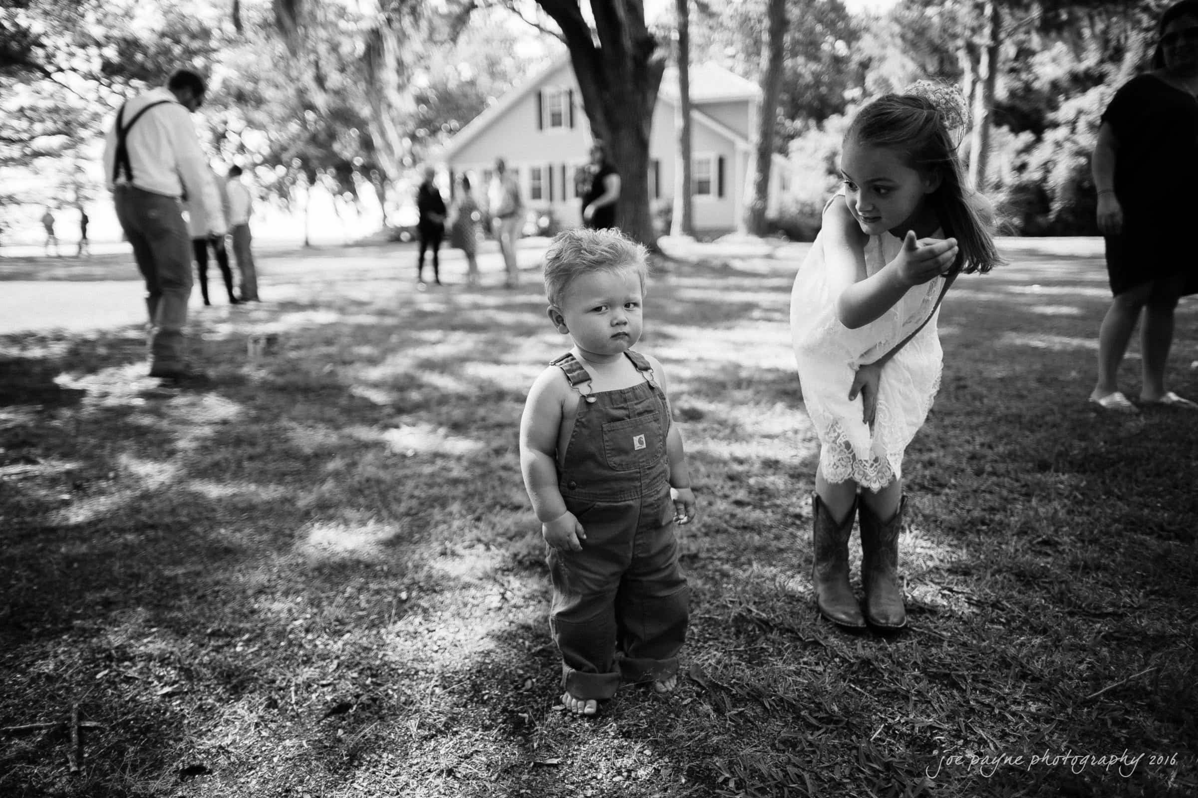 new bern wedding photographer - jessica & brinson A young child in overalls stands on grass, looking serious. A girl in a dress and boots bends down, smiling and pointing towards something. People and a house are blurred in the background, creating a sunny and lively outdoor scene.