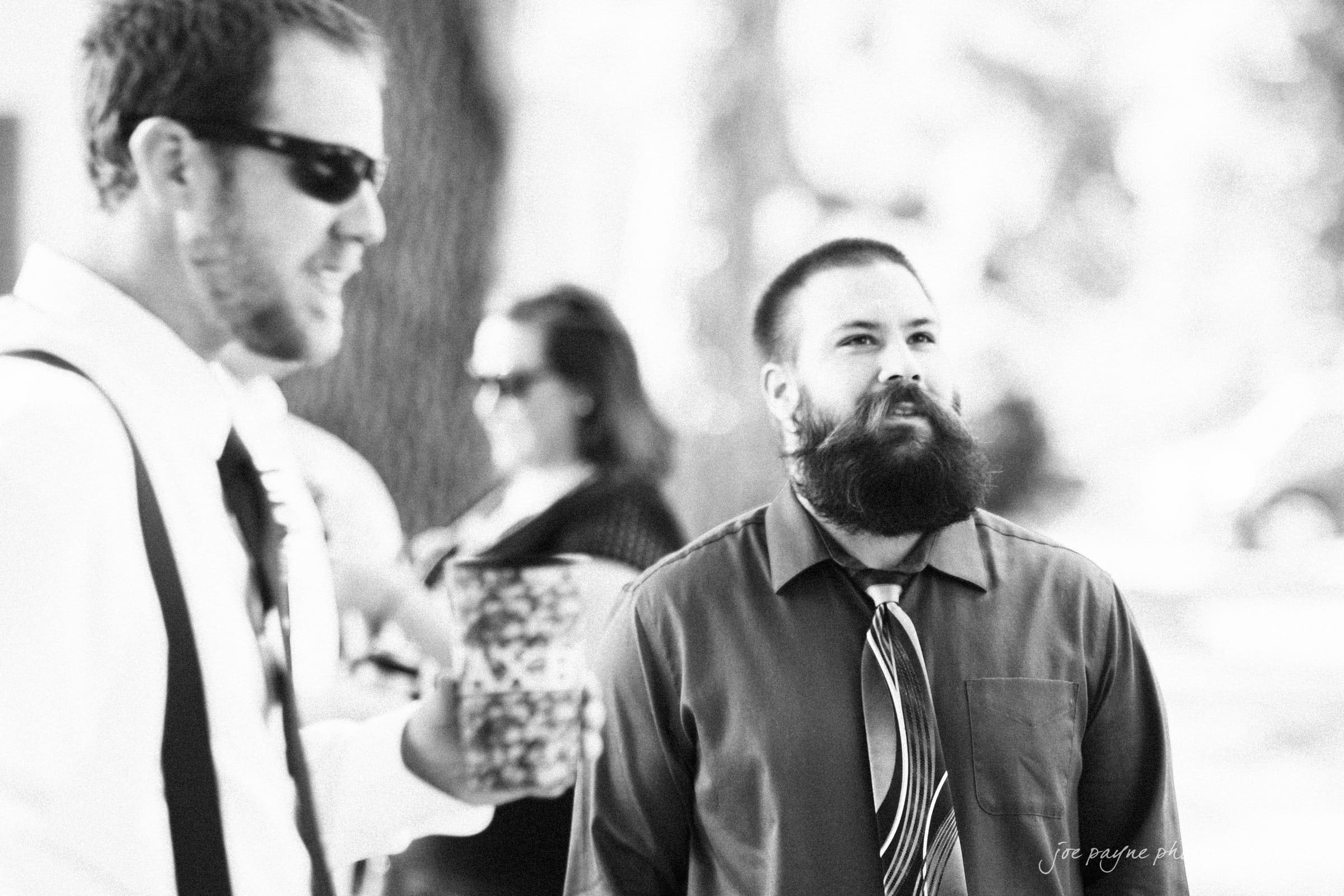 new bern wedding photographer - jessica & brinson A black-and-white photo of two bearded men outdoors. One wears sunglasses, a white shirt, and holds a cup. The other has a thick beard, and wears a dark shirt with a patterned tie. People in the background are out of focus.