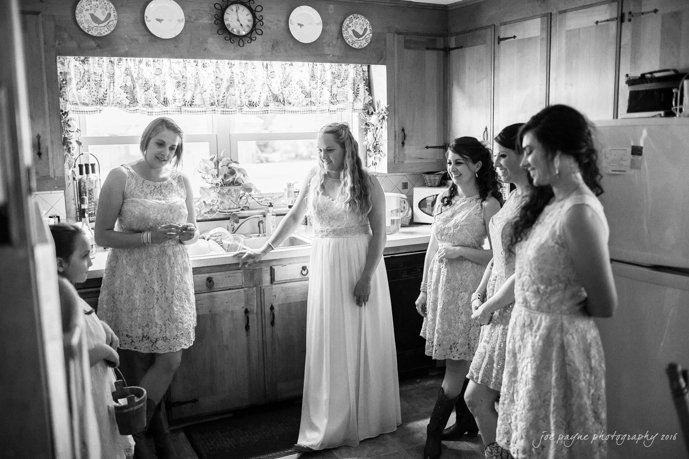 new bern wedding photographer - jessica & brinson A bride and four bridesmaids share a moment in a cozy kitchen. They are wearing matching dresses with lace details. The scene is lit by natural light coming through a window. The atmosphere is warm and joyful.