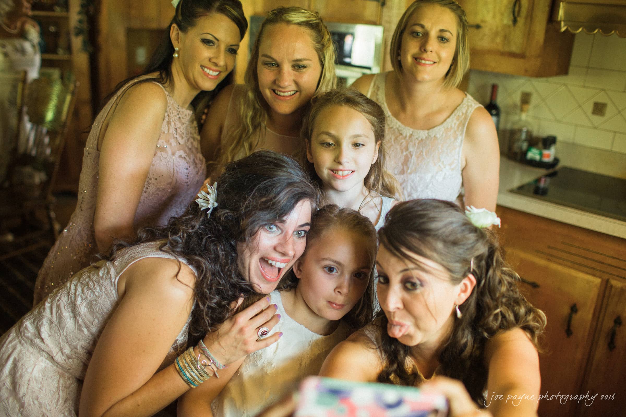 new bern wedding photographer - jessica & brinson A group of six women and girls, some wearing matching dresses, gather inside a warmly lit room. They are taking a selfie, smiling and making playful faces, capturing a joyful moment.