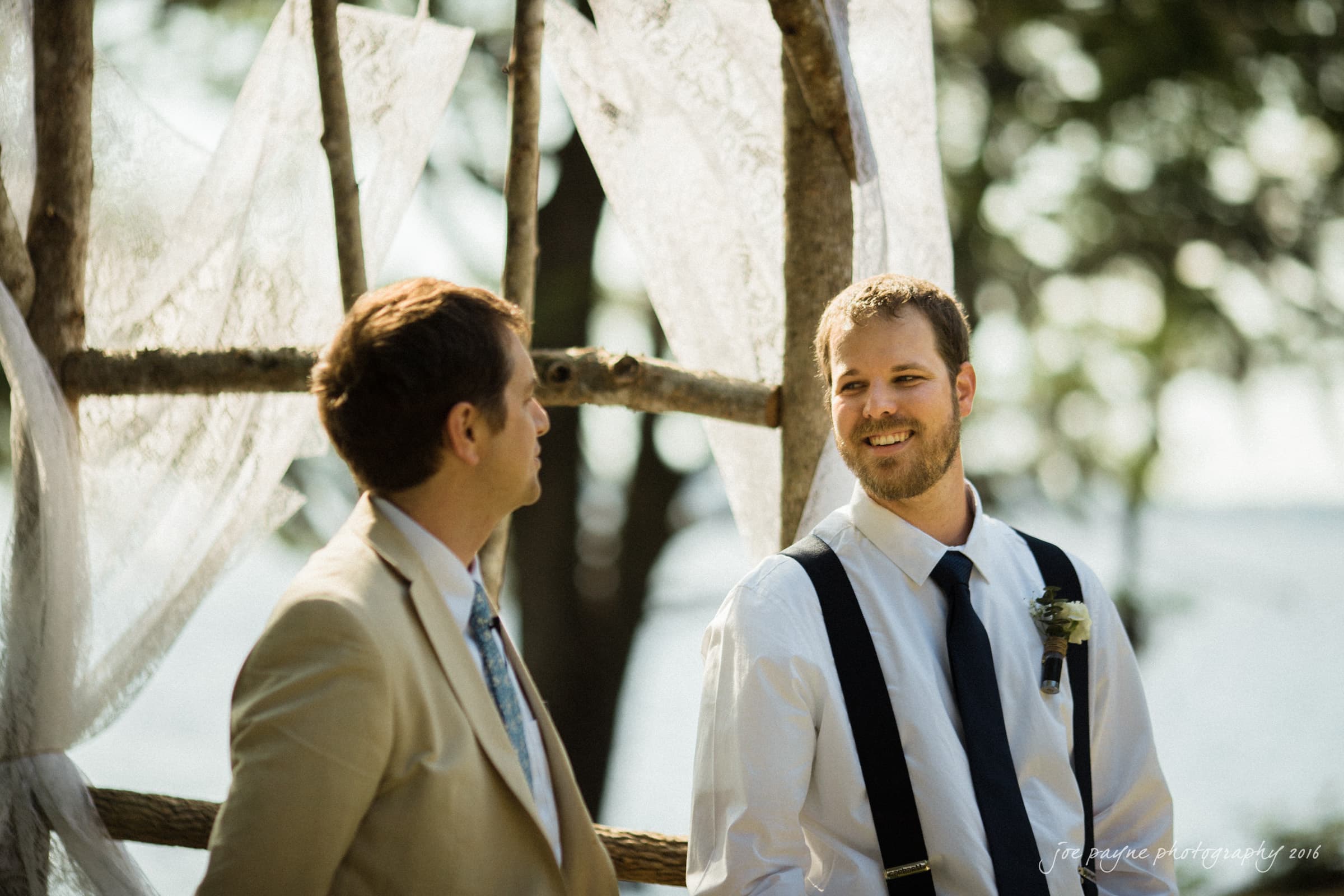 new bern wedding photographer - jessica & brinson Two men are standing outdoors, engaged in conversation. One wears a beige suit, and the other has a white shirt with suspenders and a tie. They stand by a rustic wooden arch draped with lace, with trees and water visible in the background.