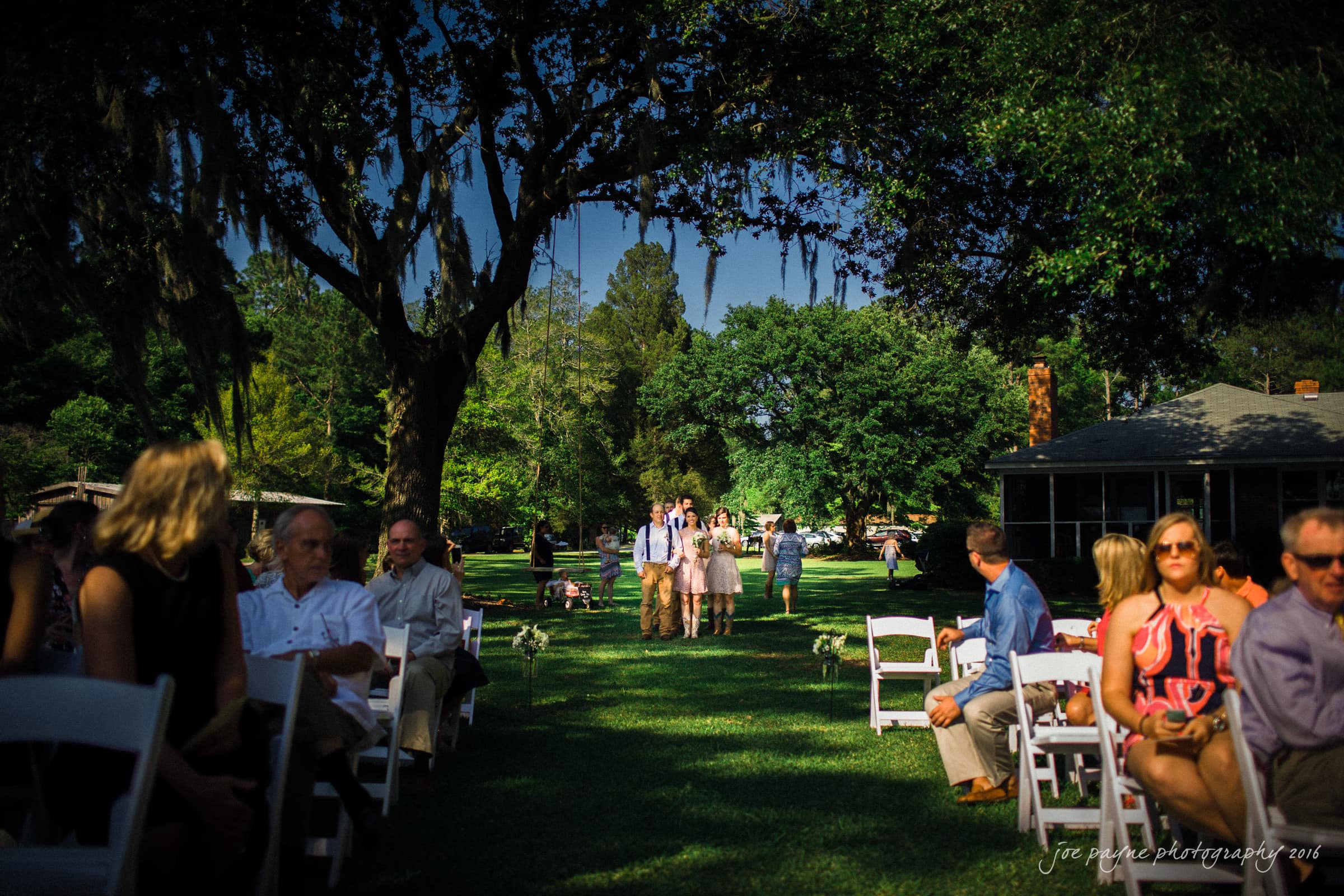 new bern wedding photographer - jessica & brinson A vibrant outdoor wedding scene with white chairs on both sides of a grassy aisle. Guests sit under lush, leafy trees, while a wedding party approaches from the distance. The sun filters through, casting dappled shadows on the ground.