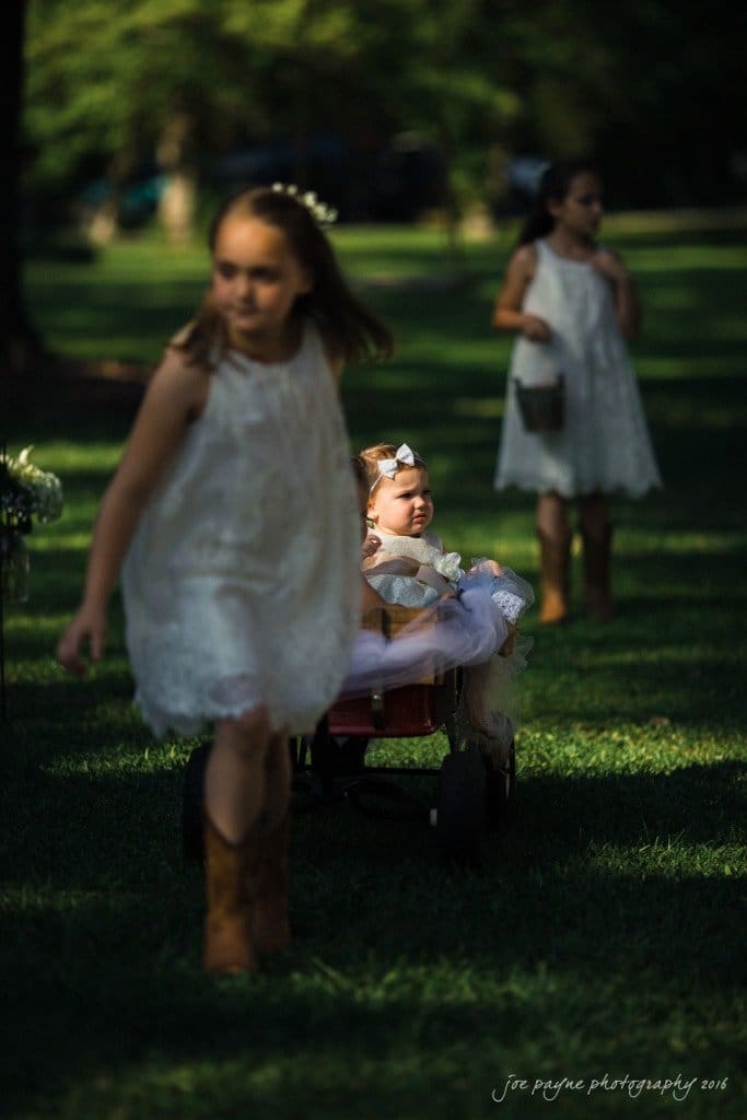 new bern wedding photographer - jessica & brinson Three young girls in white dresses walk on grass, with one girl pulling a red wagon holding a baby in a dress and bow. Sunlight creates dappled shadows on the scene.