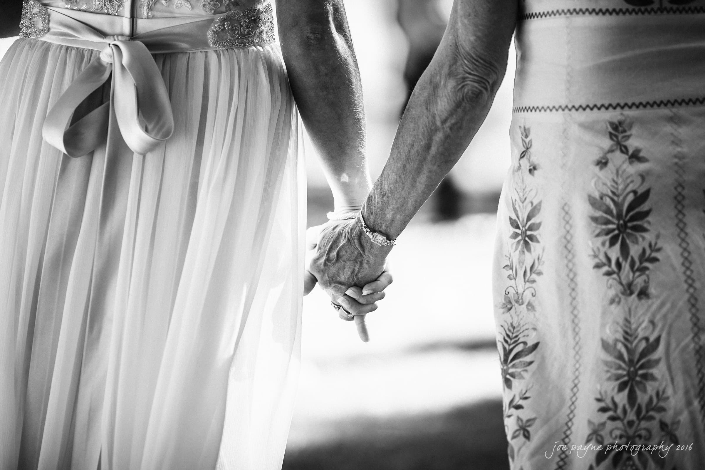 new bern wedding photographer - jessica & brinson A black and white image of two people holding hands. One wears a flowing dress with a ribbon tie; the other wears a patterned dress. They stand outdoors, suggesting a peaceful, supportive moment.