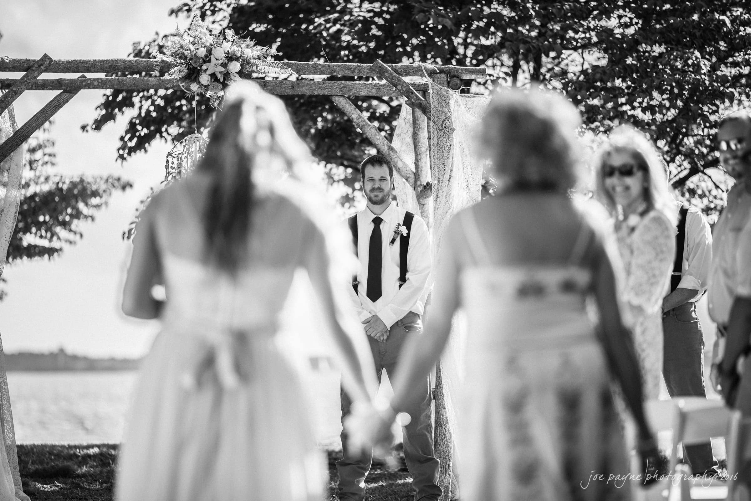 new bern wedding photographer - jessica & brinson A black-and-white photo of a bride walking down the aisle with her parents at an outdoor wedding. The groom, dressed in a shirt and suspenders, smiles under a wooden arch adorned with flowers. Family and friends watch in the sunlight.