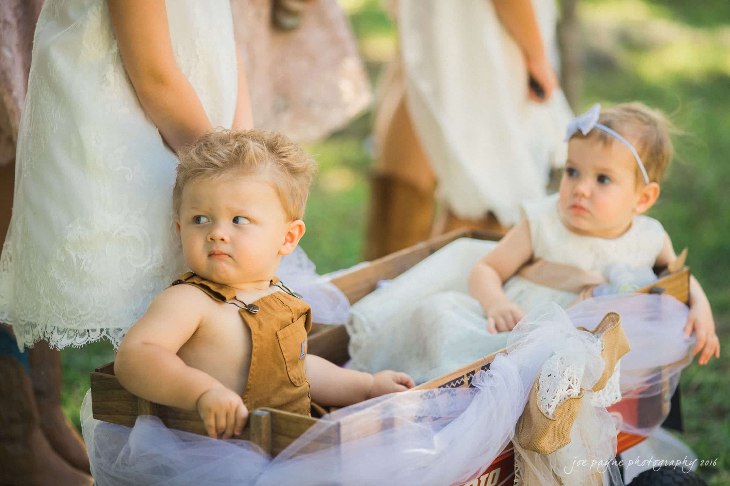 new bern wedding photographer - jessica & brinson Two toddlers are sitting in a wooden wagon decorated with white tulle. The boy is wearing brown overalls, and the girl is in a white dress with a bow headband. They appear outdoors on a sunny day.