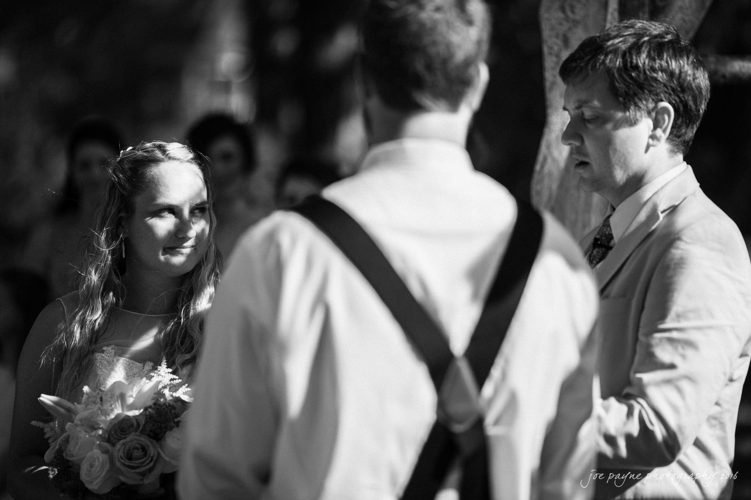 new bern wedding photographer - jessica & brinson A black and white photo captures a wedding ceremony. A bride holds a bouquet, looking at a man in suspenders, while another man in a suit speaks. The background is softly focused, suggesting an outdoor setting with guests.