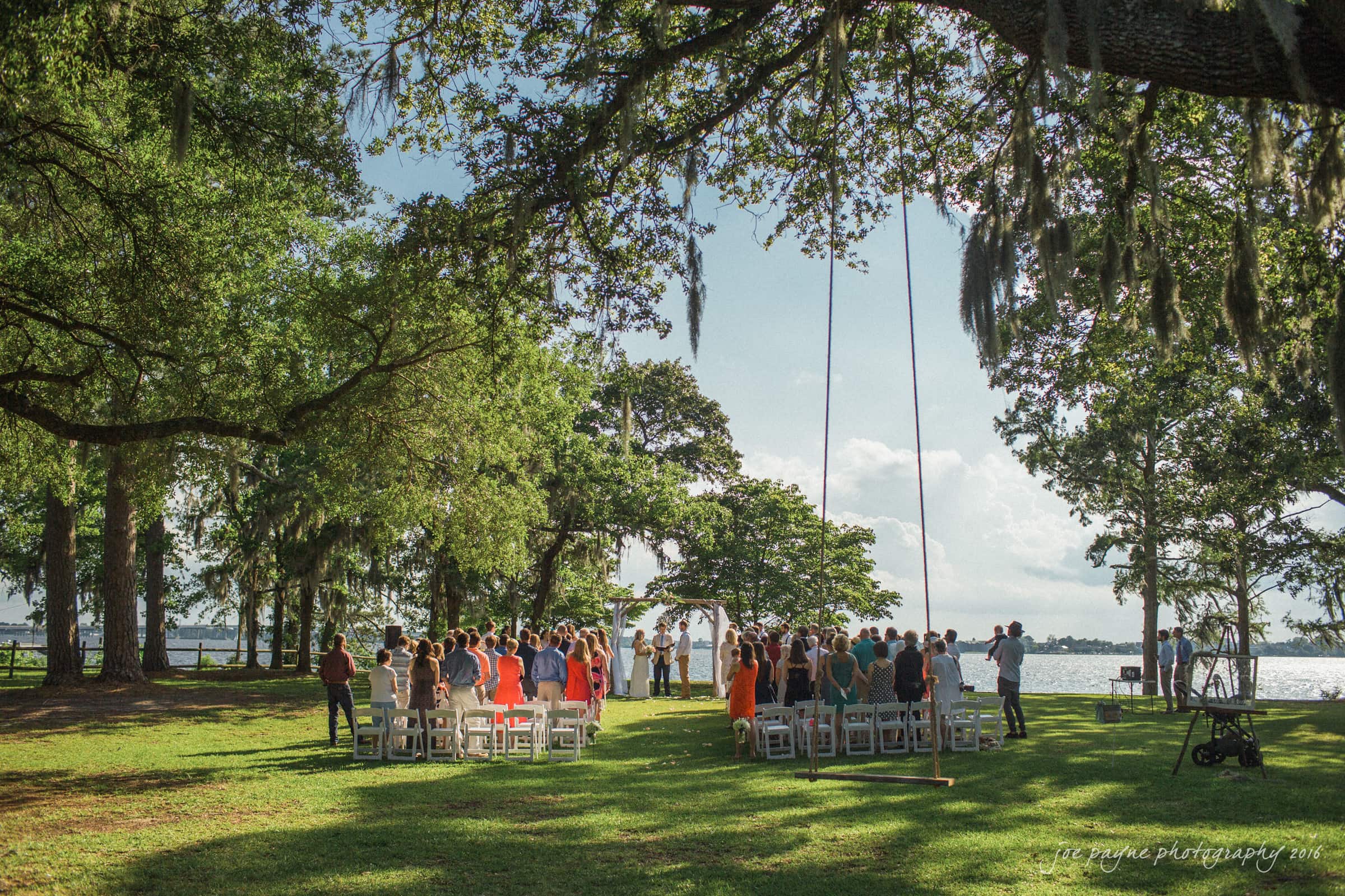new bern wedding photographer - jessica & brinson A serene outdoor wedding ceremony under large oak trees with moss. Guests are seated on white chairs facing a couple at an arch near a lake. The sky is blue and sunlit, casting light shadows on the grassy area.