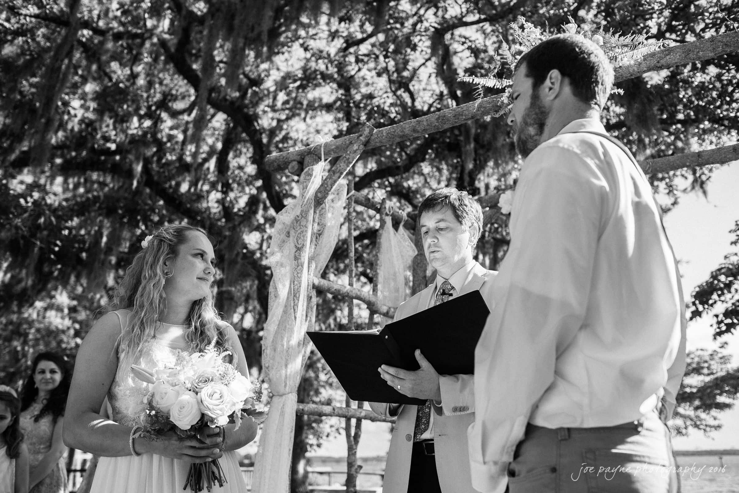 new bern wedding photographer - jessica & brinson A black-and-white photo of a wedding ceremony outdoors. A bride holding a bouquet stands facing a groom, while an officiant reads from a book. They are under a rustic arch adorned with fabric, with trees visible in the background.
