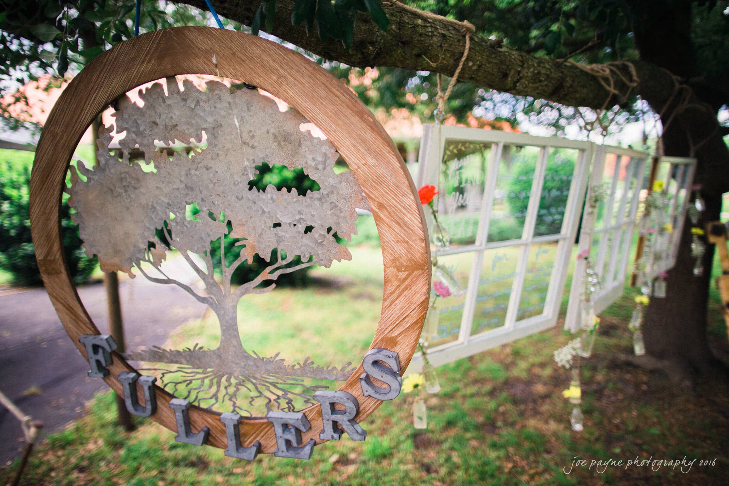 new bern wedding photographer - jessica & brinson A rustic outdoor scene with a wooden sign depicting a large tree and the word FULLERS hanging from a branch. In the background, vintage window frames and flowers are strung along the same branch. Lush greenery surrounds the setup.