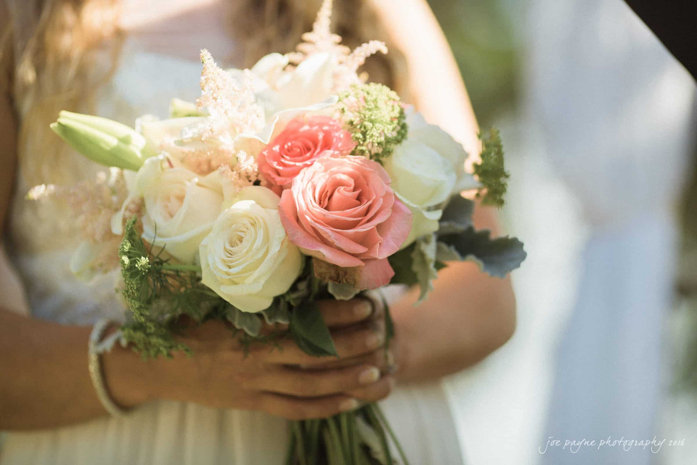 new bern wedding photographer - jessica & brinson A close-up of a bridal bouquet featuring roses in shades of pink and white, surrounded by greenery. The bouquet is being held by a person in a white dress, partially visible in the background.