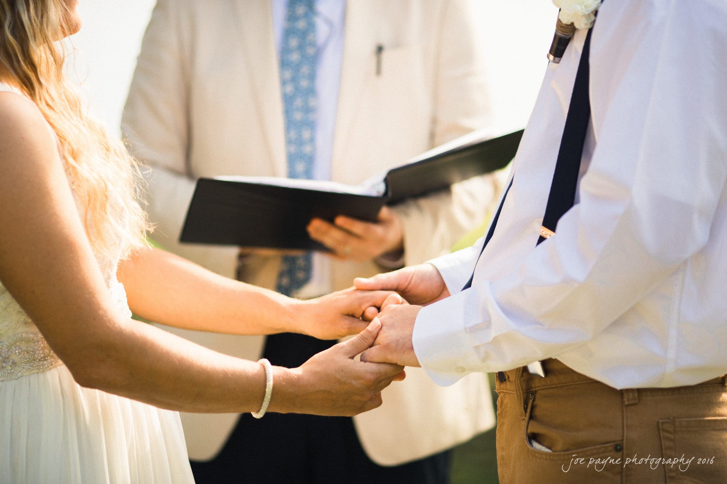 new bern wedding photographer - jessica & brinson A bride and groom stand holding hands during a wedding ceremony. The bride is wearing a white dress, and the groom is dressed in a white shirt and brown pants. An officiant holding a book stands in the background. The setting is outdoors.