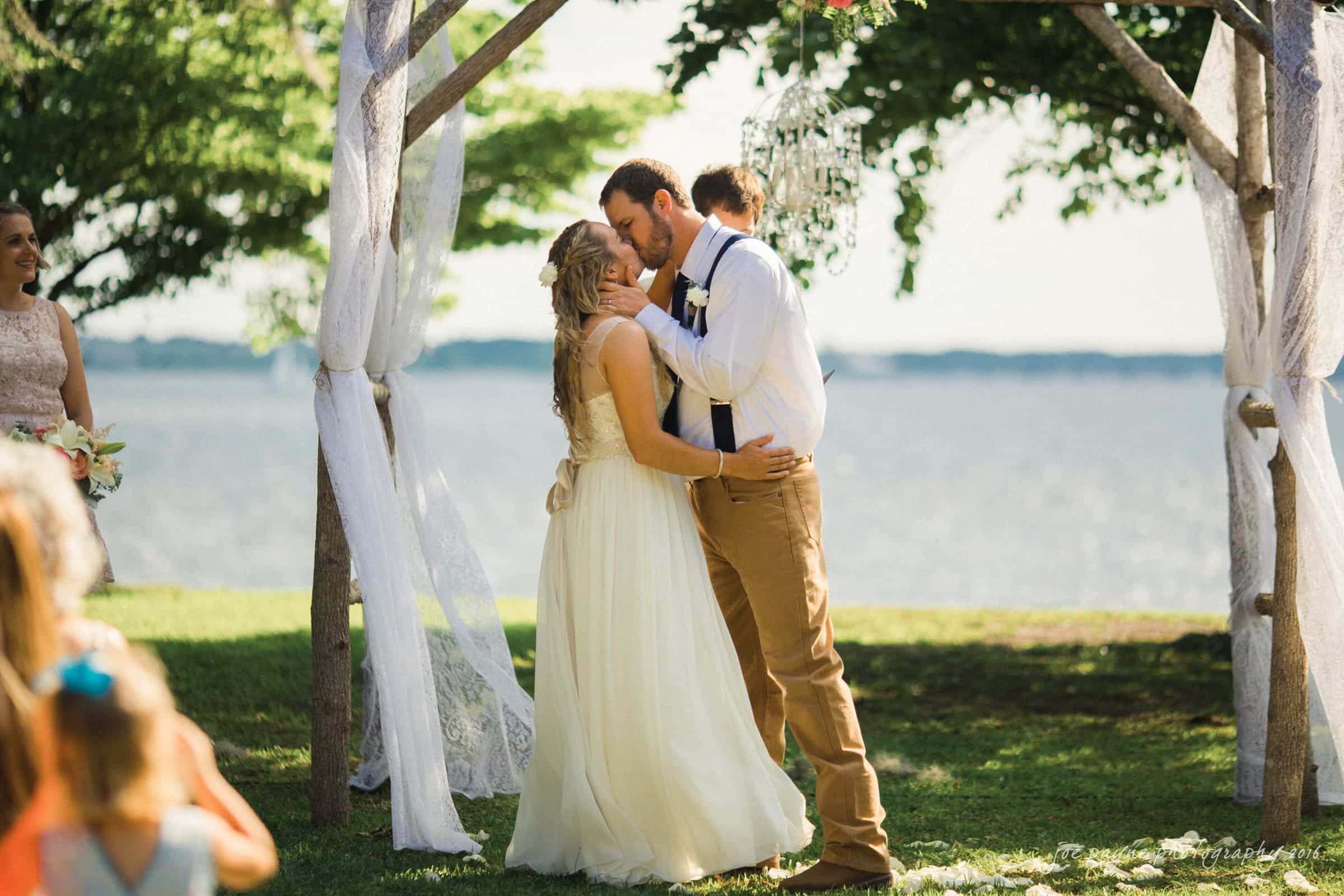 new bern wedding photographer - jessica & brinson A bride and groom share their first kiss under a rustic wooden arch draped with white fabric during an outdoor wedding ceremony by a lake. Green grass and shade from trees surround the sunny scene. Spectators are partially visible in the foreground.